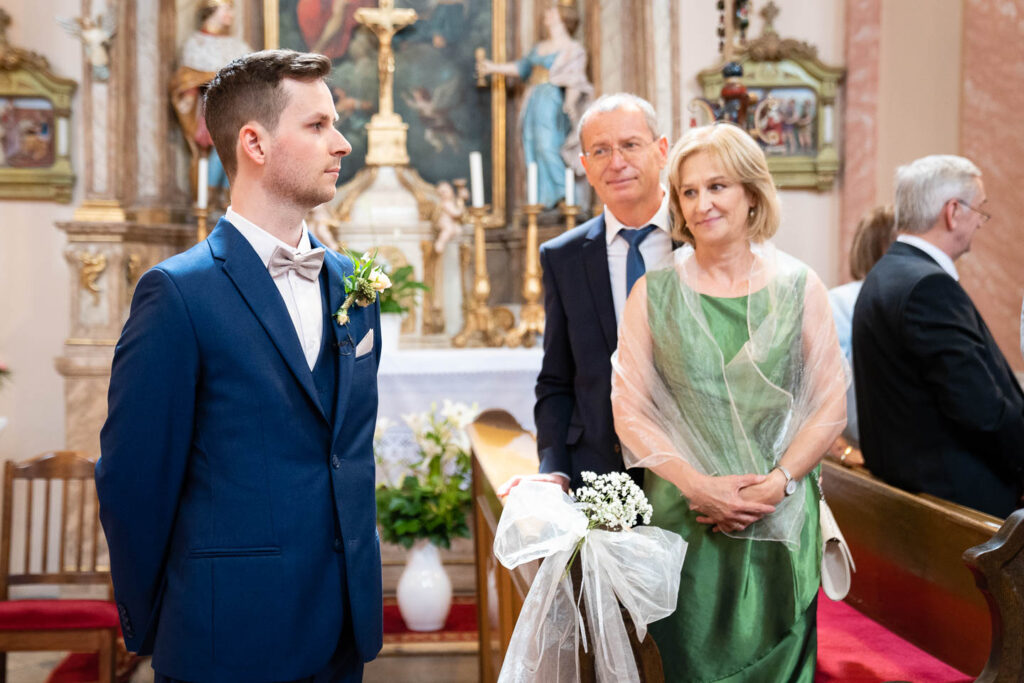 Der Bräutigam wartet in der Kirche auf die Braut, während ihre Eltern zuschauen. Erster Blick. Berührend. Hochzeitsfotograf Wien