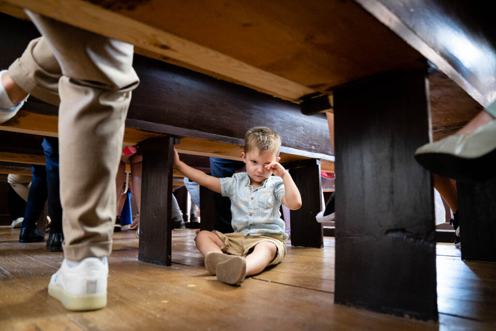 Der kleine Junge langweilte sich unter den Kirchenbänken bei der kirchlichen Trauung. Besonderes eingefangenes Momentfoto. Hochzeitsfotograf Wien
